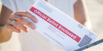 A young woman places her absentee voter ballot for the 2020 presidential election into a blue United States Postal mailbox.