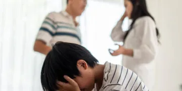 Asian boy kid sitting and crying on bed while parents having fighting or quarrel conflict at home. Child covering face and eyes with hands do not want to see the violence. Domestic problem in family.