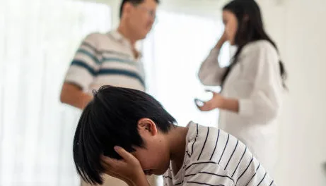 Asian boy kid sitting and crying on bed while parents having fighting or quarrel conflict at home. Child covering face and eyes with hands do not want to see the violence. Domestic problem in family.